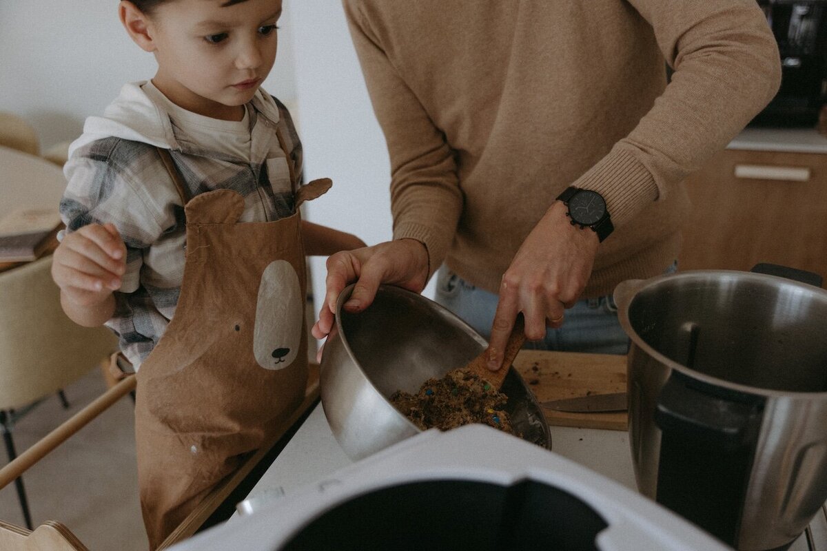 Le père cuisine avec son fils qui porte un tablier ourson. Il lui montre comment verser la pâte dans un plat. Séance famille par Kamescop, photographe en Loire-Atlantique.