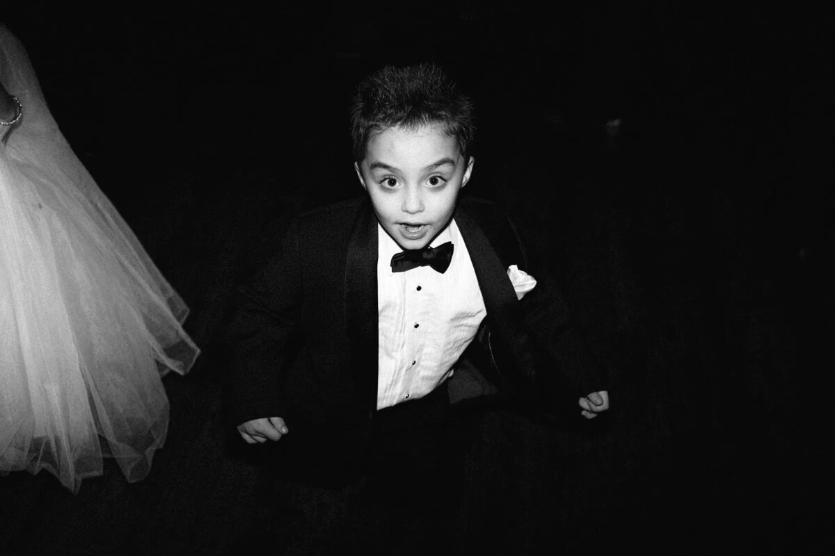 A young boy in a tuxedo and bow tie looks up at the camera with a surprised expression; to the left, part of a person in a white dress is visible—captured candidly by a film photographer NJ against a dark background.