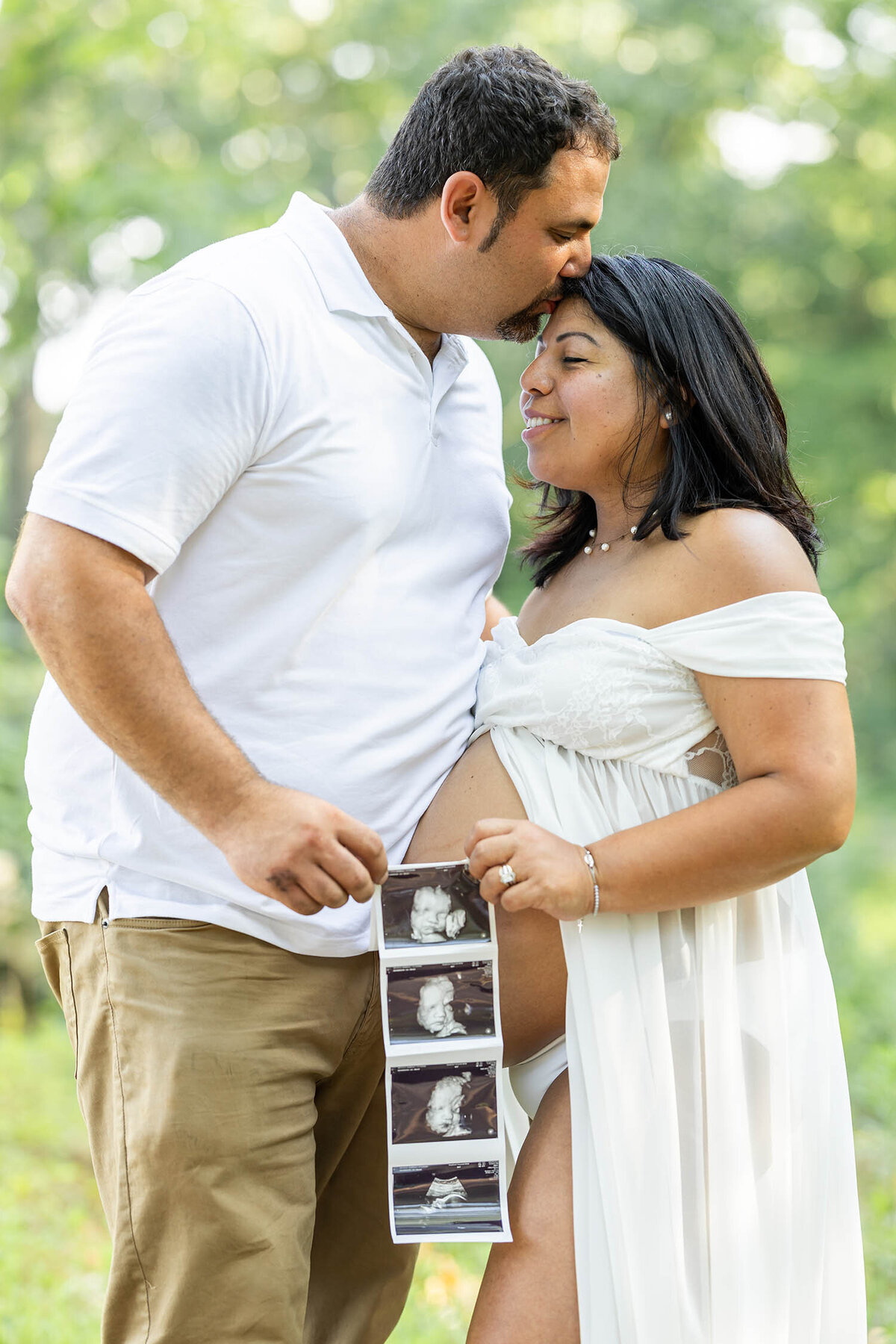A man wearing a white shirt and tan pants kisses a pregnant woman on her forehead as they both stand holding a baby sonogram during their maternity session in Connecticut 