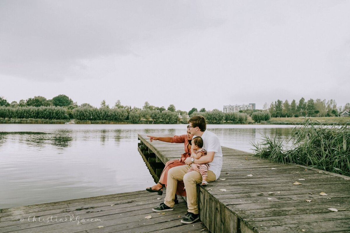 Parents sitting on a wooden dock, pointing across the calm lake while holding their baby.