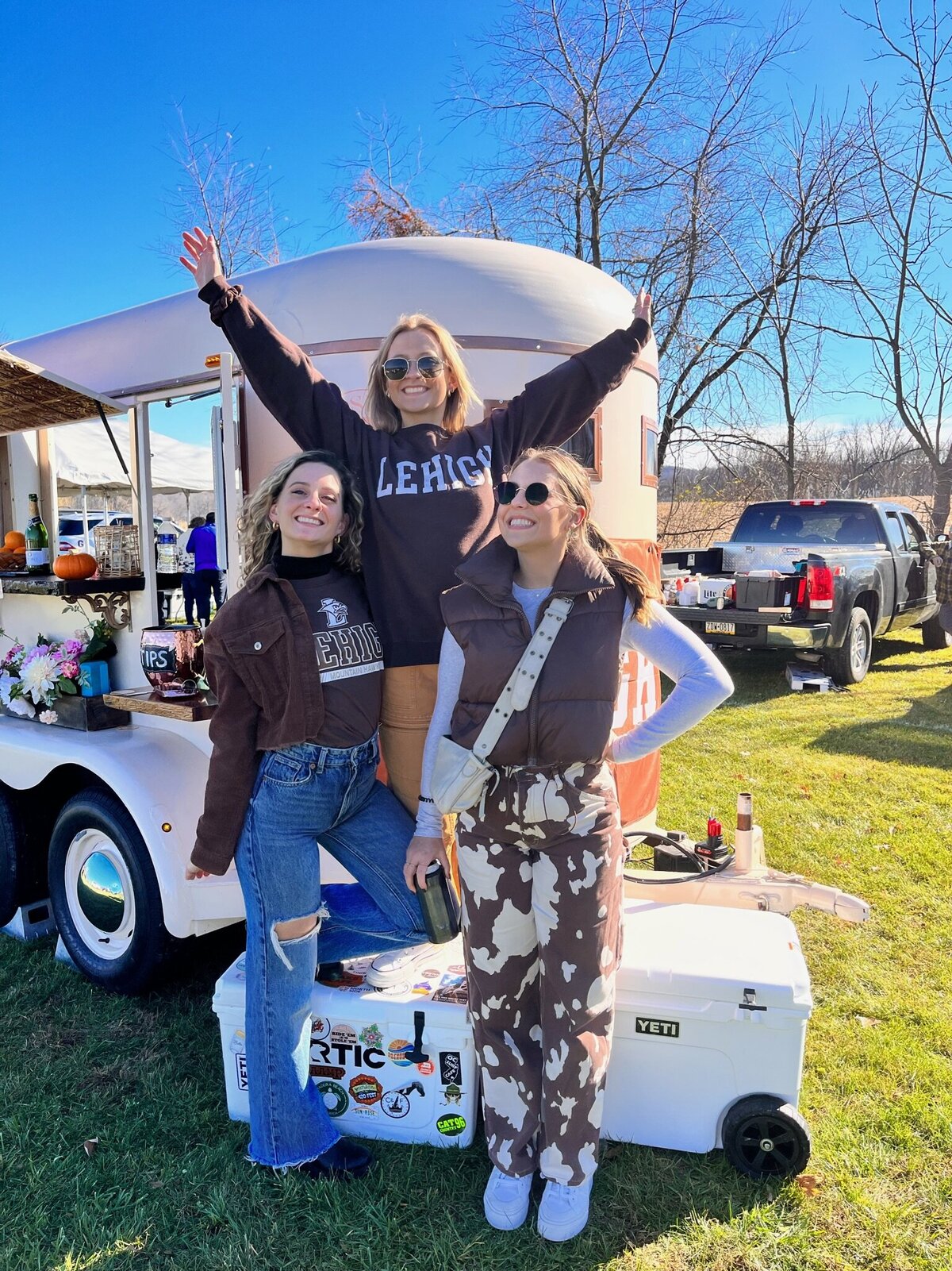 Three girls stand in front of trailer at a tailgate event