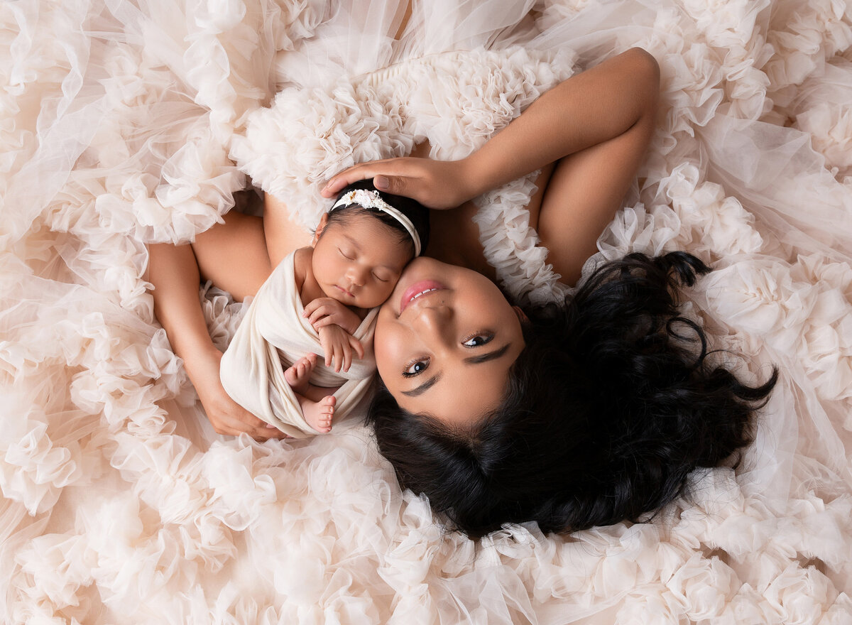 Rochel Konik Photography | Top NYC Brooklyn Newborn Photographer captures mom laying atop a flowing blush gown with her her baby resting on her shoulder. Aerial image.