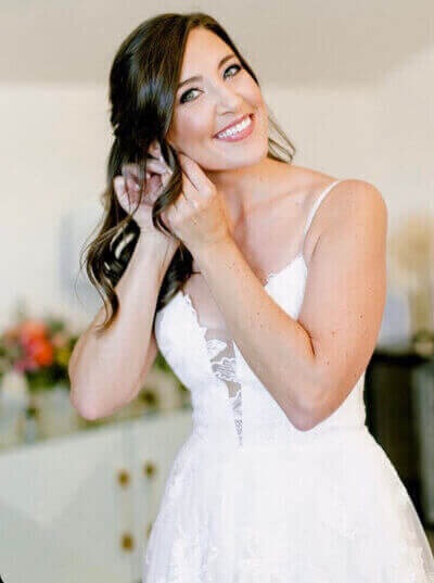 Bride with soft waves and radiant makeup putting on earrings, featuring elegant bridal hair and makeup services by an expert makeup artist for wedding day prep