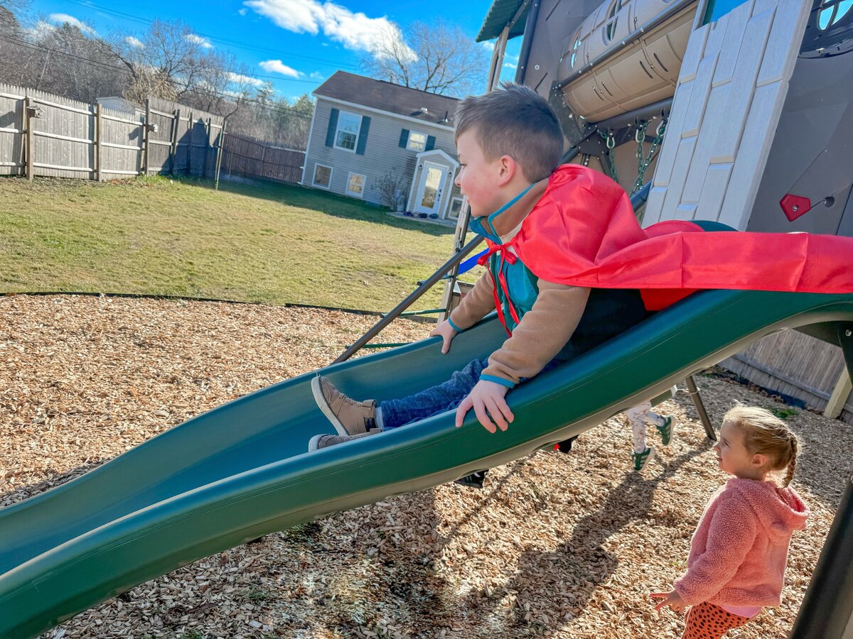 Student going down the slide in the cape on the playground at House of Parker Preschool in Gorham Maine where we believe that play isn’t just something children do—it’s how they learn. Through play, children discover the world around them, express their creativity, build relationships, and develop the foundational skills they’ll carry with them for life.