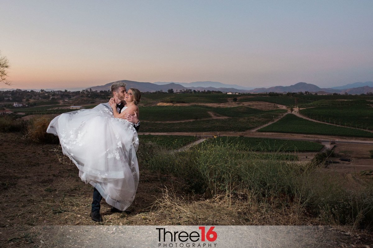 Groom lifts his Bride up and kisses her cheek on a hilltop overlooking the Temecula Valley