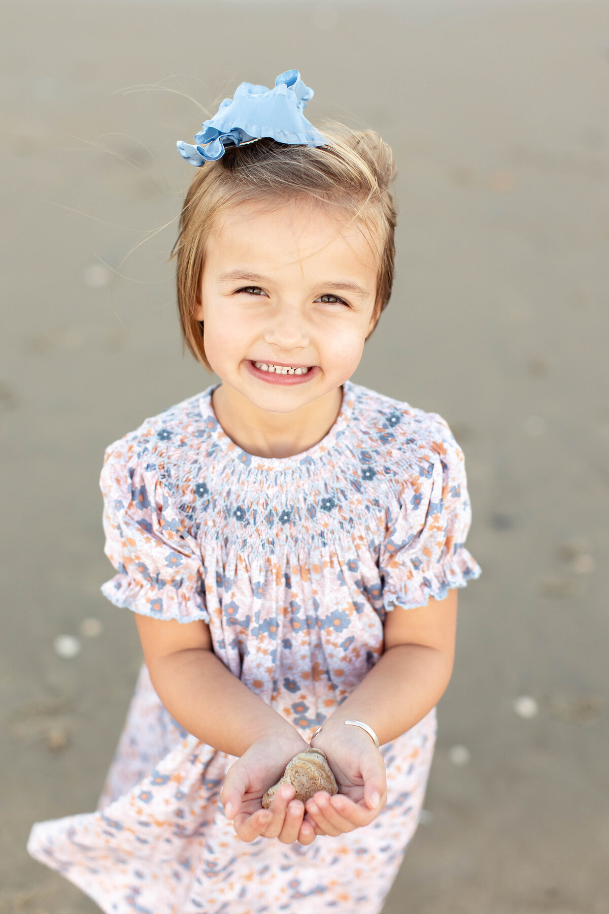 A young girl dressed in a pink smocked dress and blue bow smiles as she stands on the sandy shore in Virginia Beach, holding out a shell during her Brooke Tucker photography session.
