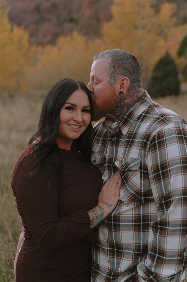 A man kisses his wife's forehead as she looks at the camera and smiles. It's fall time as they're standing outside somewhere in Idaho Falls.