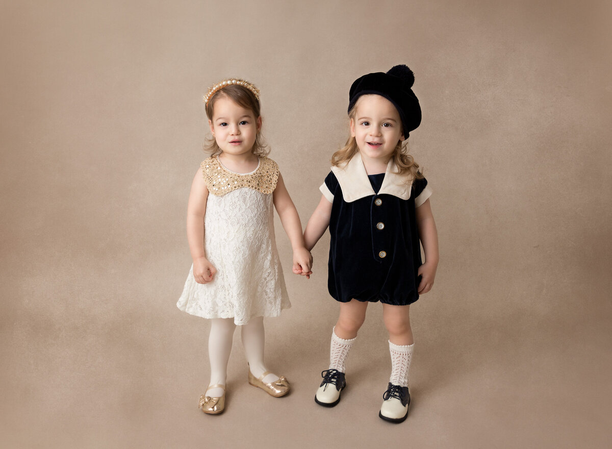 Toddler siblings are standing holding hands and smiling at the camera.