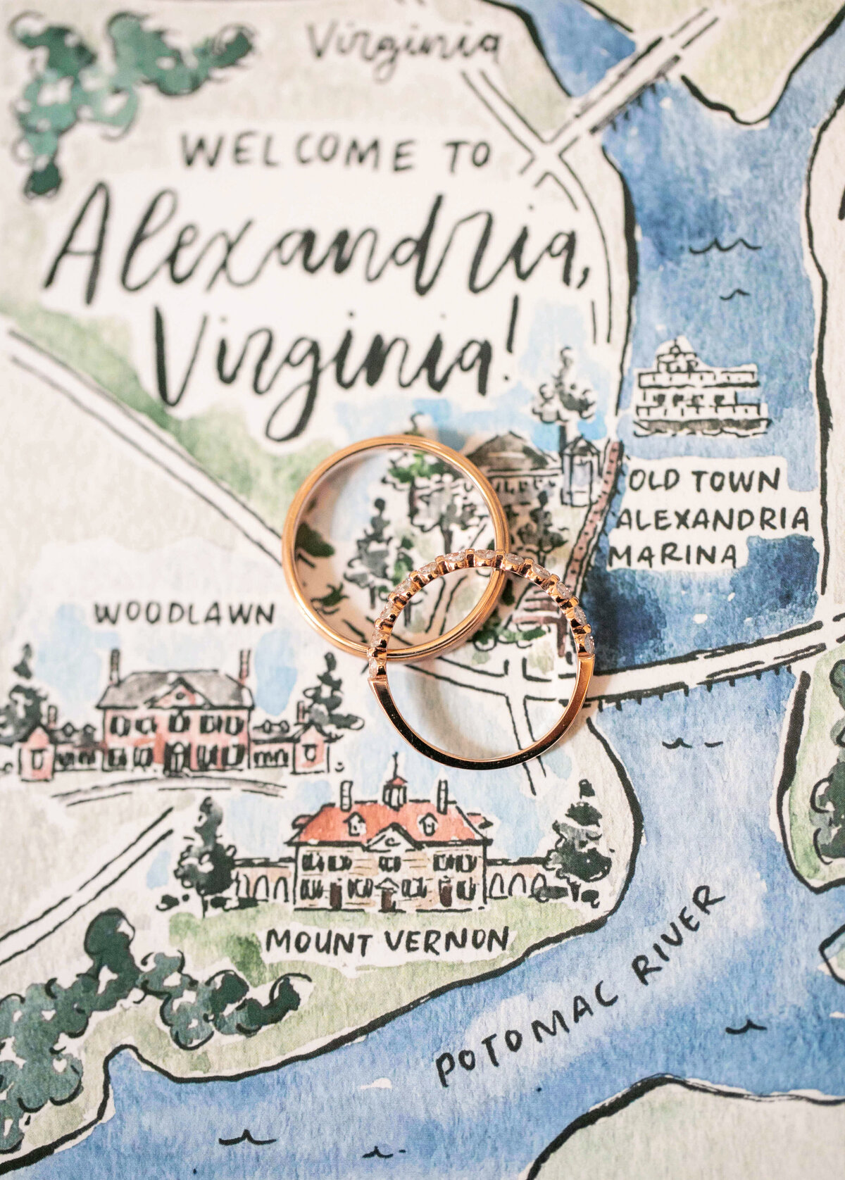 A hand-drawn map illustration featuring Alexandria, Virginia, highlighting locations like Old Town and Mount Vernon along the Potomac River. Two gold rings are placed on top of the map, symbolizing a wedding or engagement.