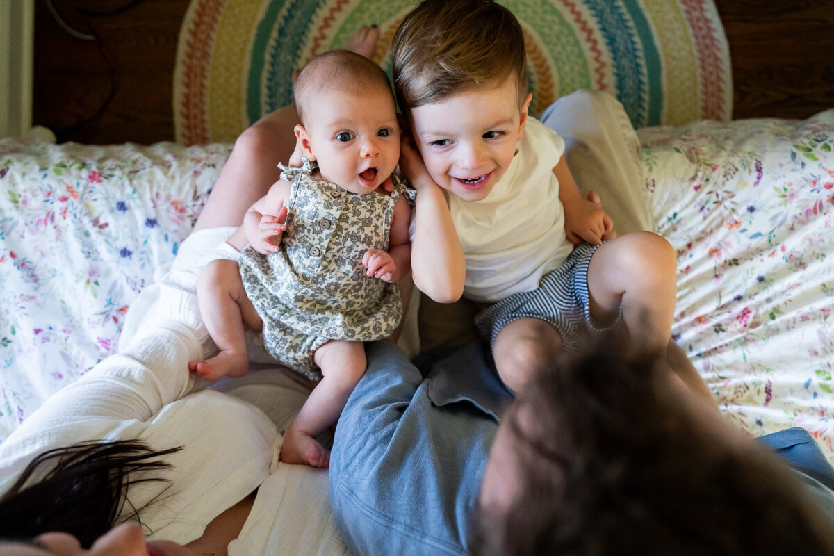 overhead photo of newborn baby girl and toddler brother being held by parents on laps at edge of bed at home