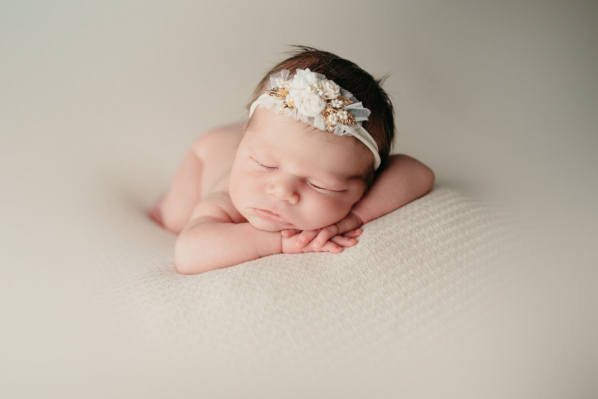 Newborn girl nude lying on tummy resting cheek and chin on her hands wearing white flower headband laying on white fabric backdrop