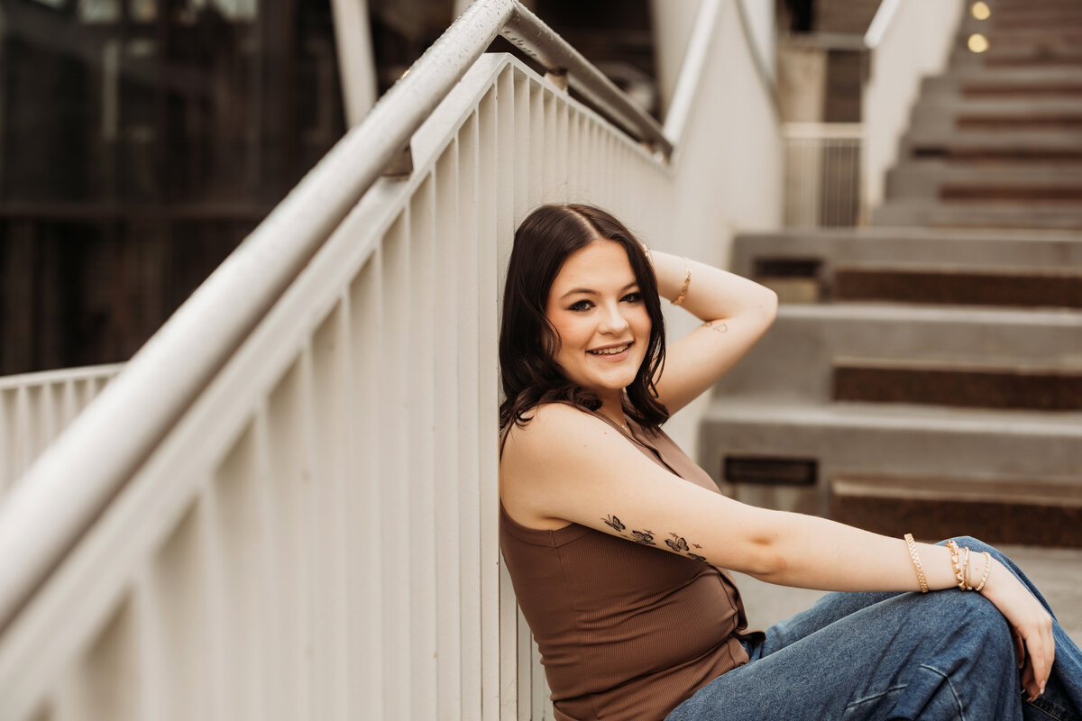 high school senior girl leans against white metal stairs in a brown tank top in downtown Denver for senior photos