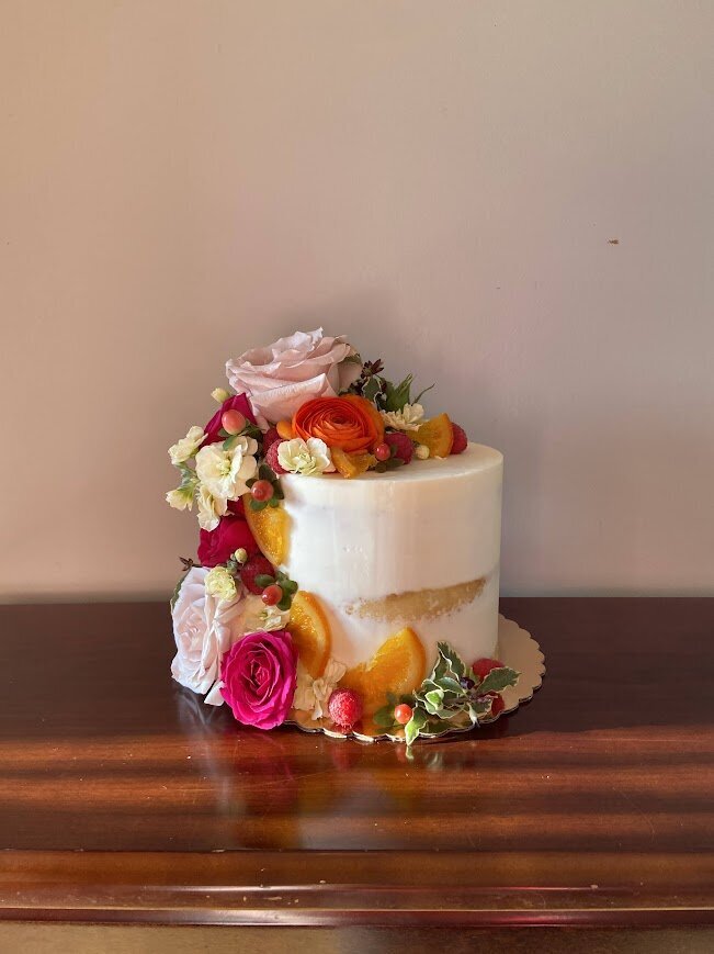 A naked style cutting cake with a cascade down the side of fresh flowers in orange, yellow, deep pink, and pale pink, with fresh raspberries and candied oranges.