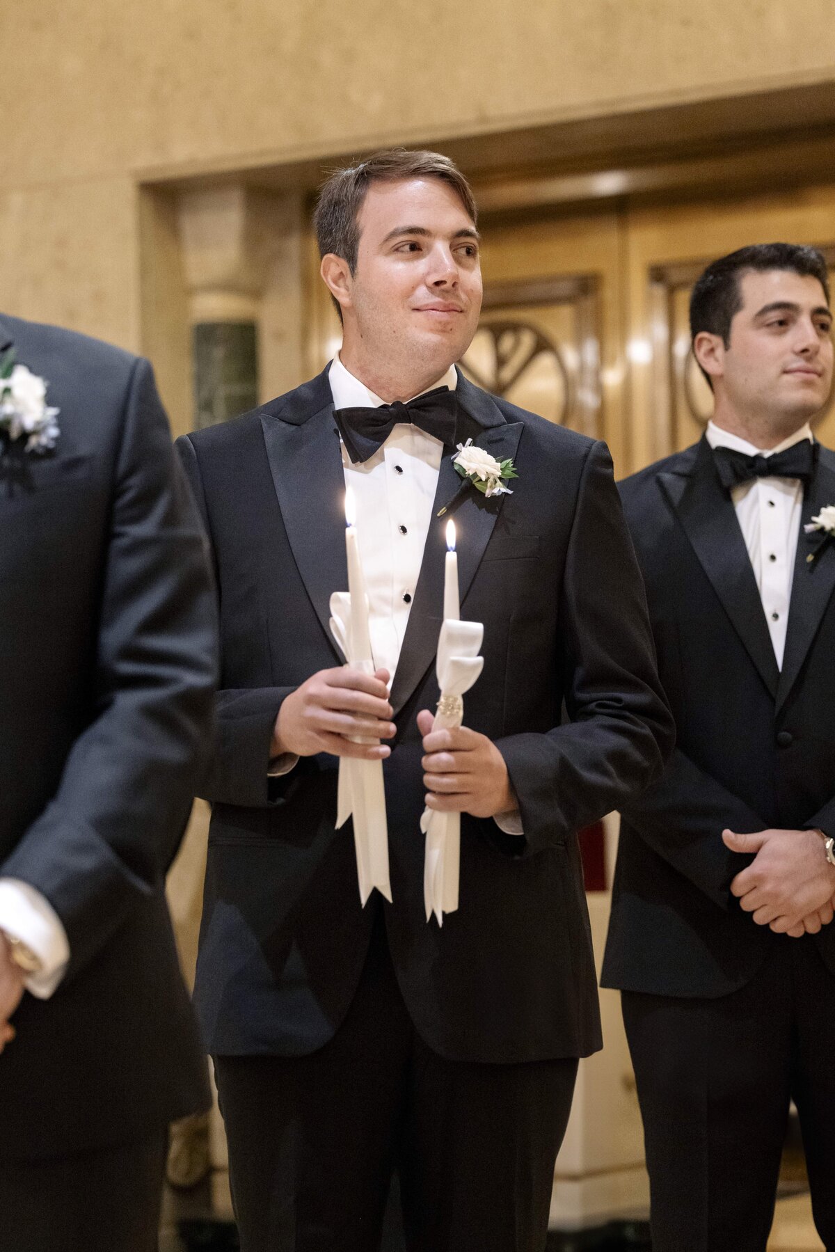 A man in a tuxedo holds two lit candles, standing indoors. He is smiling slightly and wearing a bow tie, with a boutonniere on his lapel. Another man in a tuxedo stands nearby, partially visible in the background.