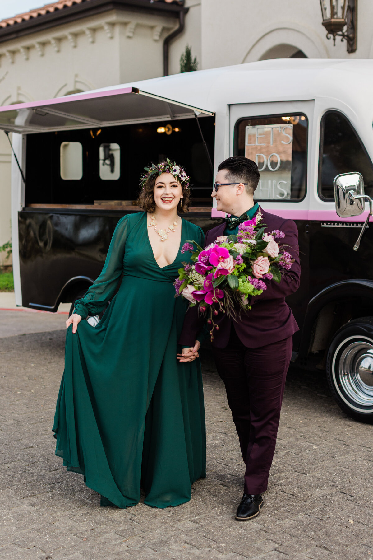 Two brides posing together in front of a food truck on their wedding day in Dallas, Texas. The bride on the left is wearing a flowing green dress with a flower crown, and the bride on the right is wearing a burgundy suit with a green bowtie and is holding a large bouquet. The food truck behind them has a neon sign in the window reading "Let's Do This."