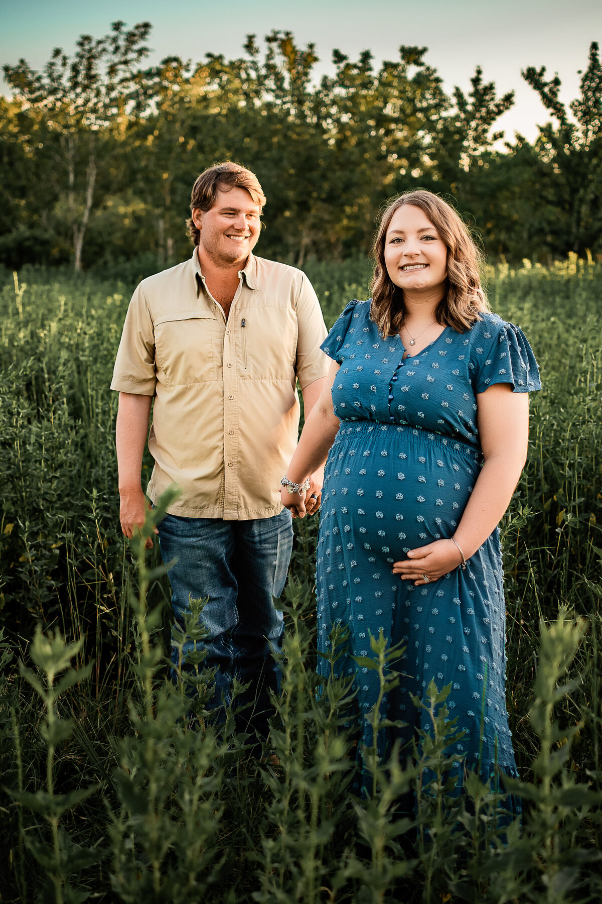 A pregnant wife leads her husband through a field of long grass as he looks at her and smiles.