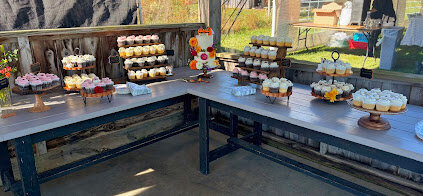 An L-shaped table display of wooden cake stands in various shapes and sizes topped with different flavors of cupcakes. At the table joining is a white two tier wedding cake decorated with fresh fall flowers. 