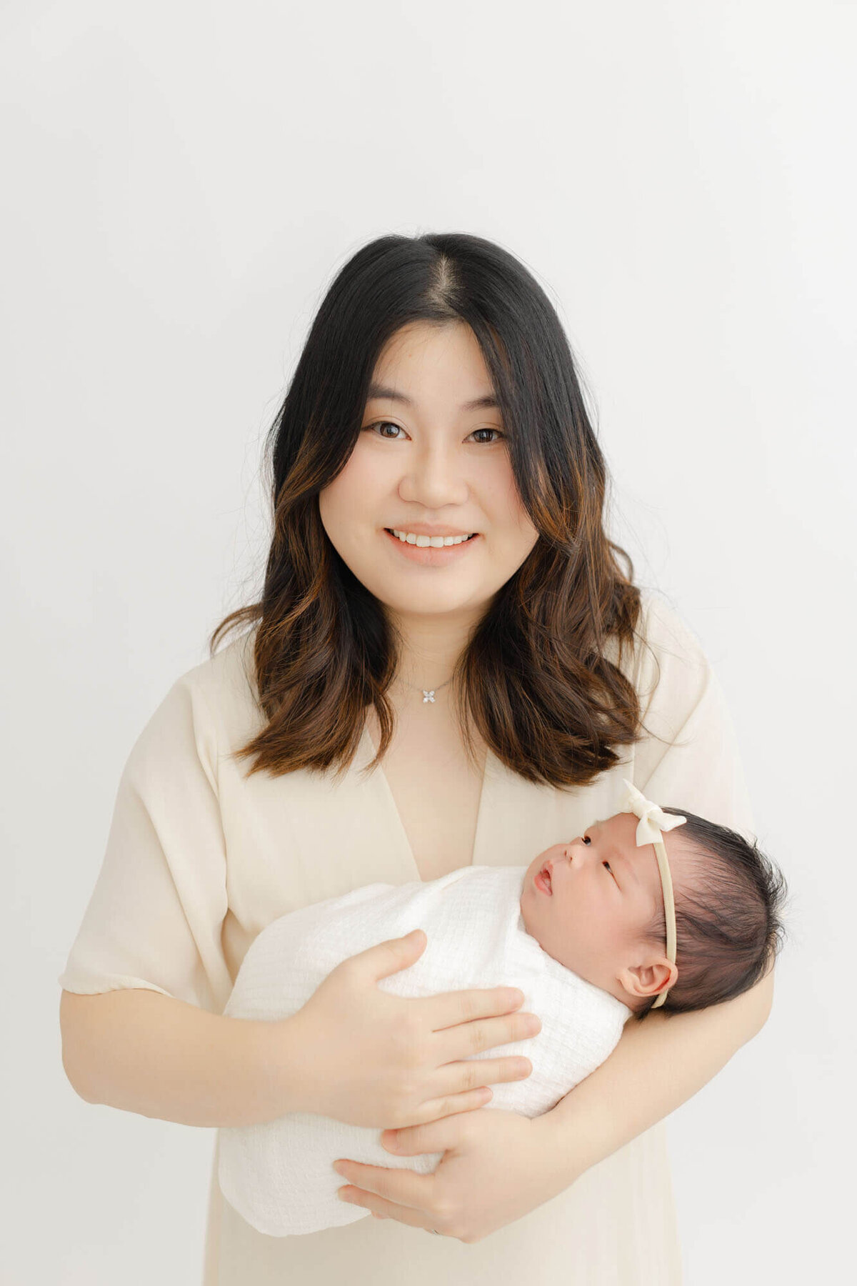 Mom dressed in a cream dress holding baby in her arms while smiling at the camera. Bab y is swaddled in white and looking up at mom.