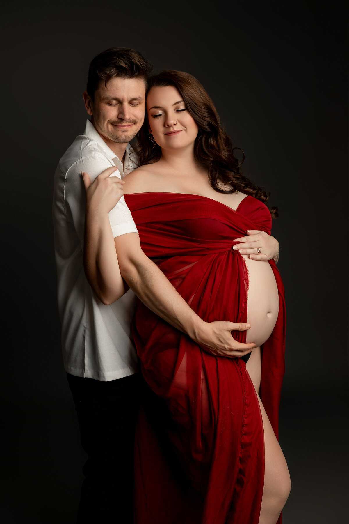 The expectant mother, in a bold red dress, stands with her husband, sharing a tender moment as they embrace their growing family. Captured by Inland Empire maternity photographer Kaitlyn Dawn Photography.