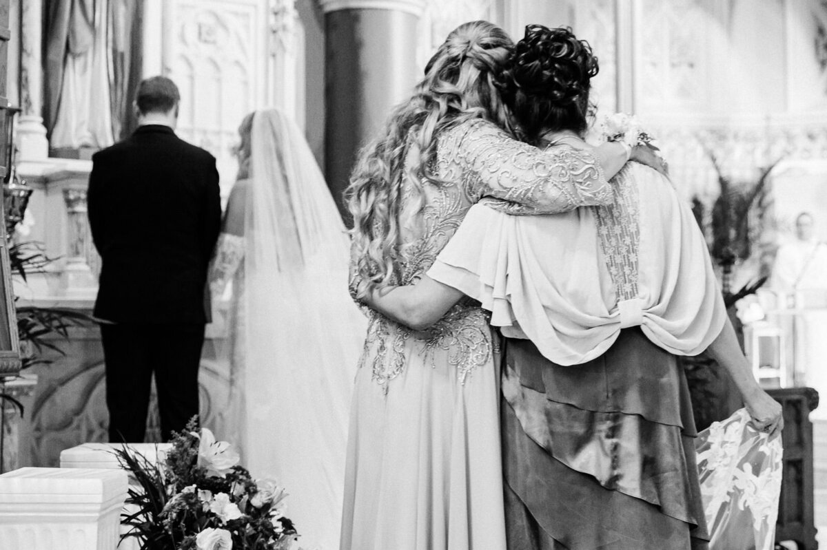 The bride’s mother and the groom’s mother sharing an emotional embrace during the wedding ceremony, captured in a candid black and white moment.