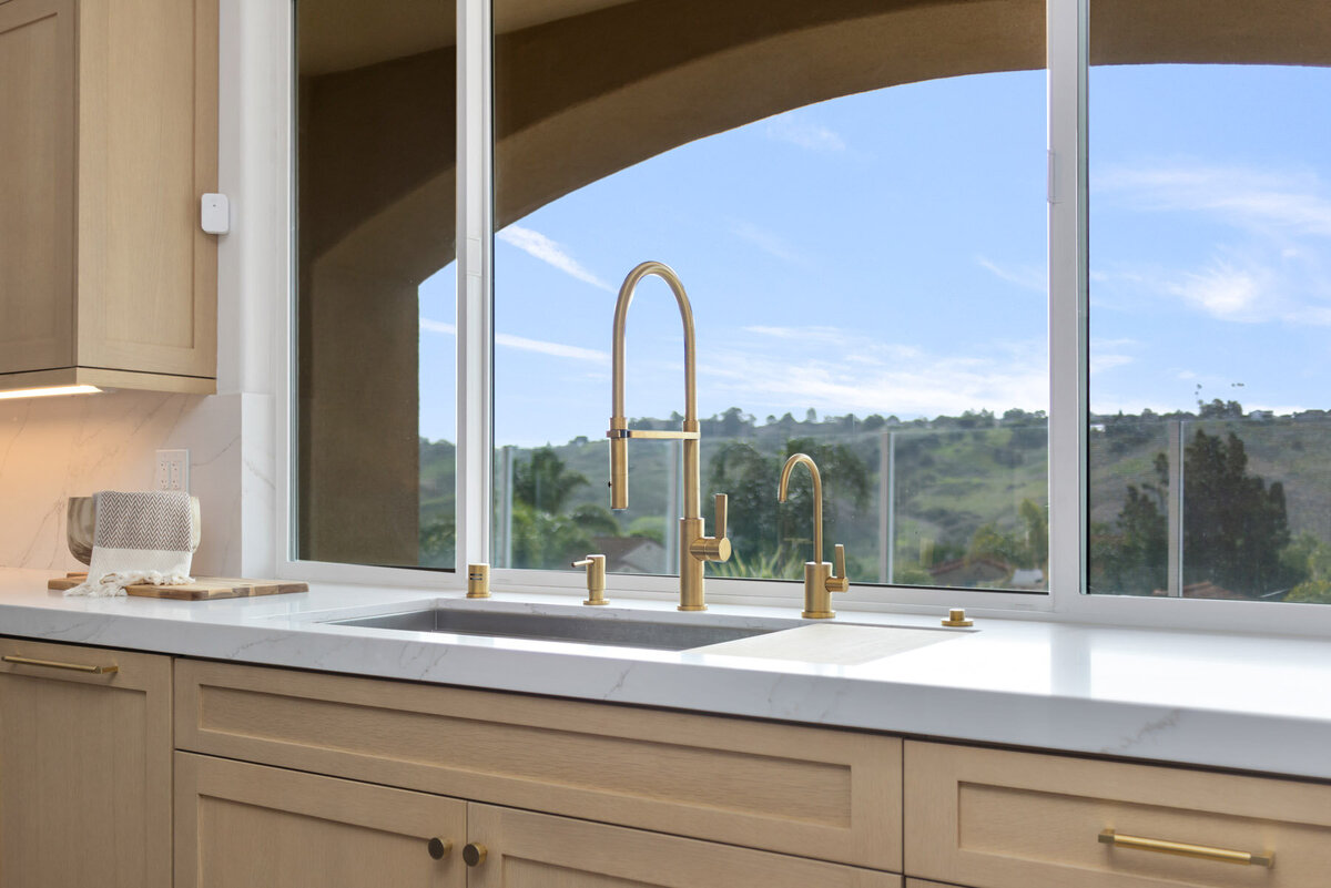 Bathed in natural light, this kitchen corner effortlessly combines luxury with practicality. The expansive arched window provides a picturesque view of the rolling hills and blue skies, acting as nature's own artwork. Golden fixtures, from the sink taps to the cabinet handles, introduce a touch of opulence, while the pristine marble surface and the patterned holder reflect an eye for detail.