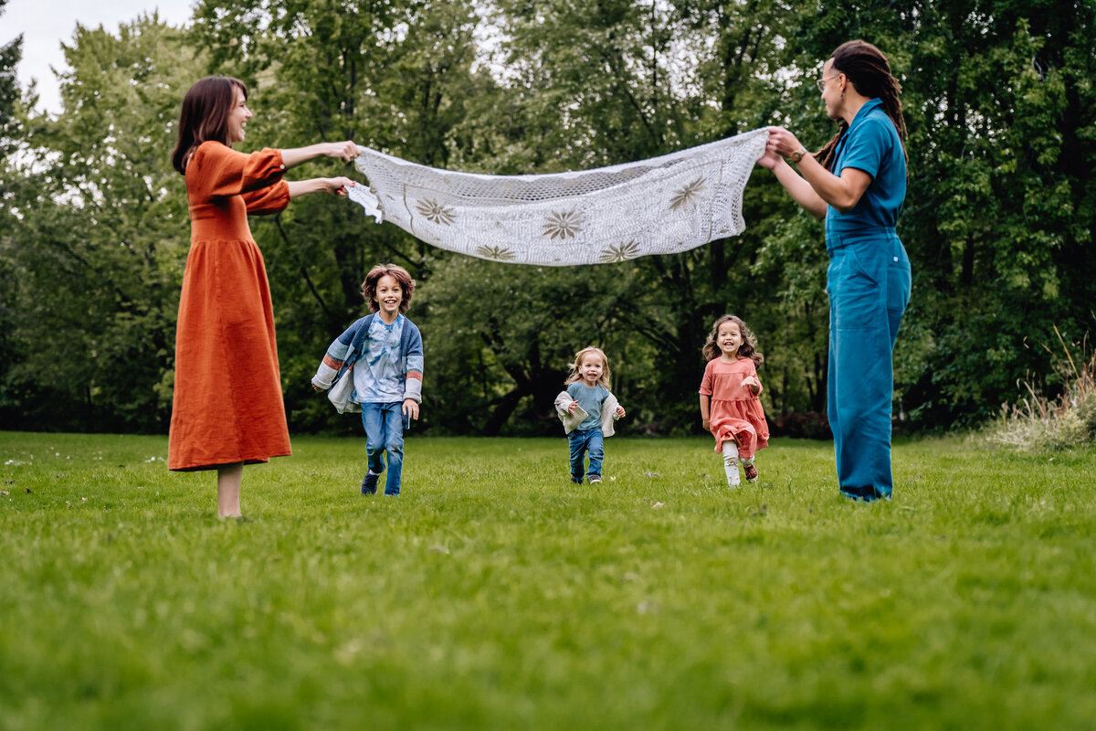 Two parents hold up a blanket while their kids run underneath.