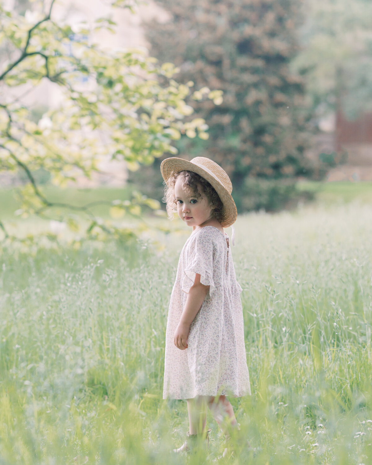 Little girl in a straw hat and dress standing in tall grass looking towrads the camera by Savannah Family photographer Courtney Cronin