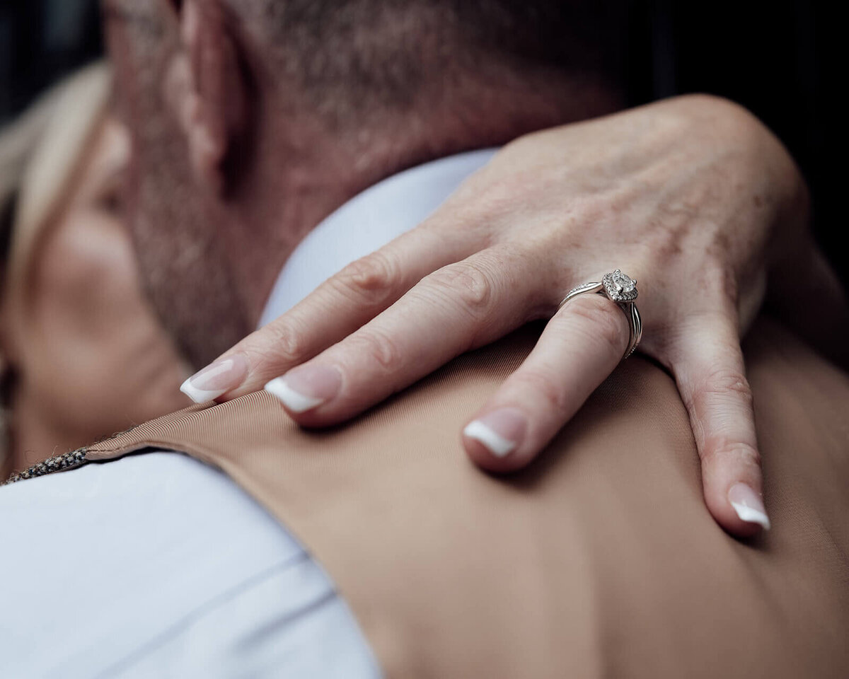 The left hand of a bride with a large diamond on her finger around the back of the grooms neck.
