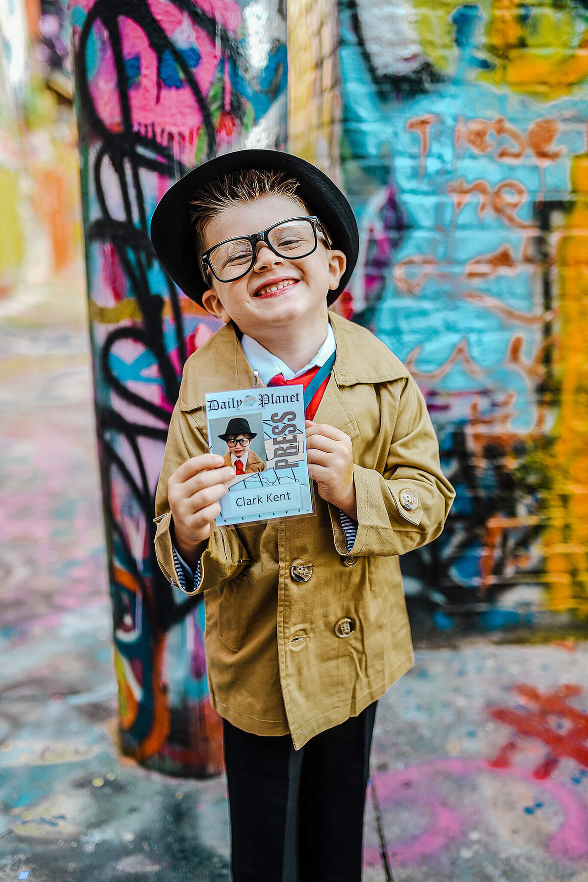 Little Boy wearing a black fedora and trench coat holding a Clark Kent press badge in graffiti alley Baltimore Maryland near MICA