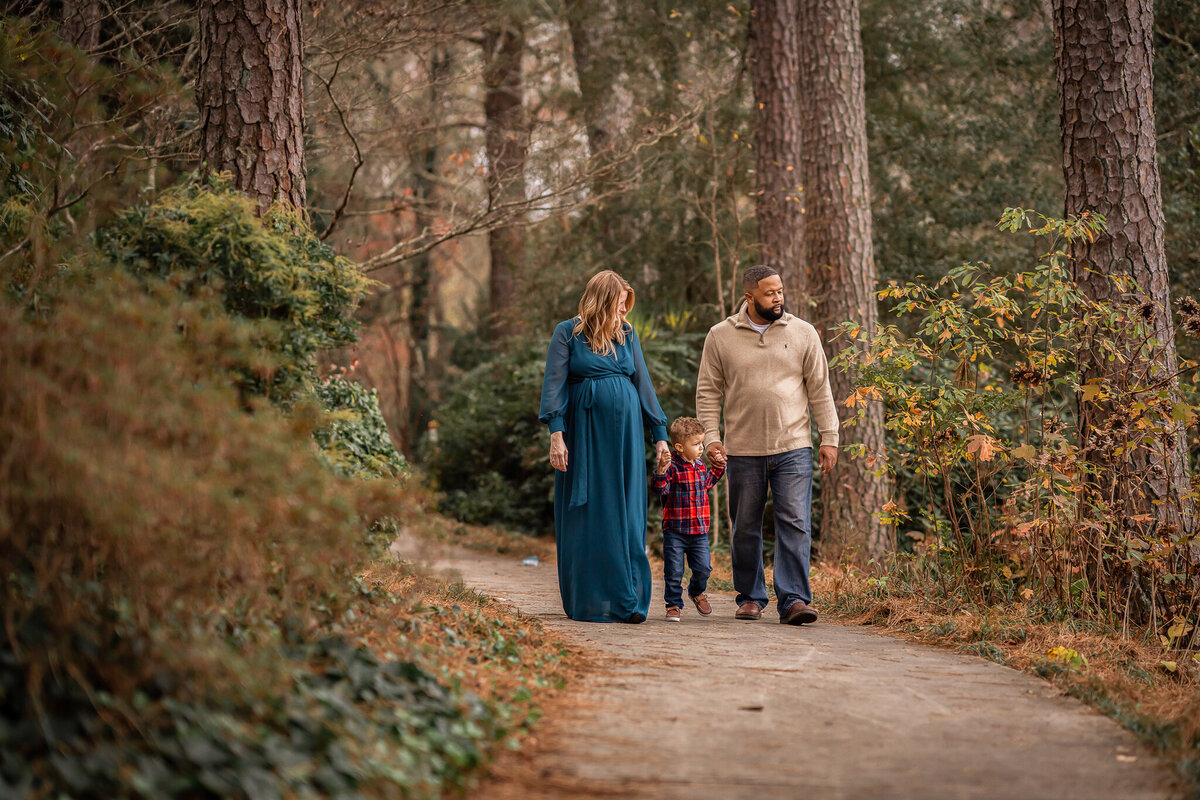 A family of three is holding hands and walking through a wooded area.
