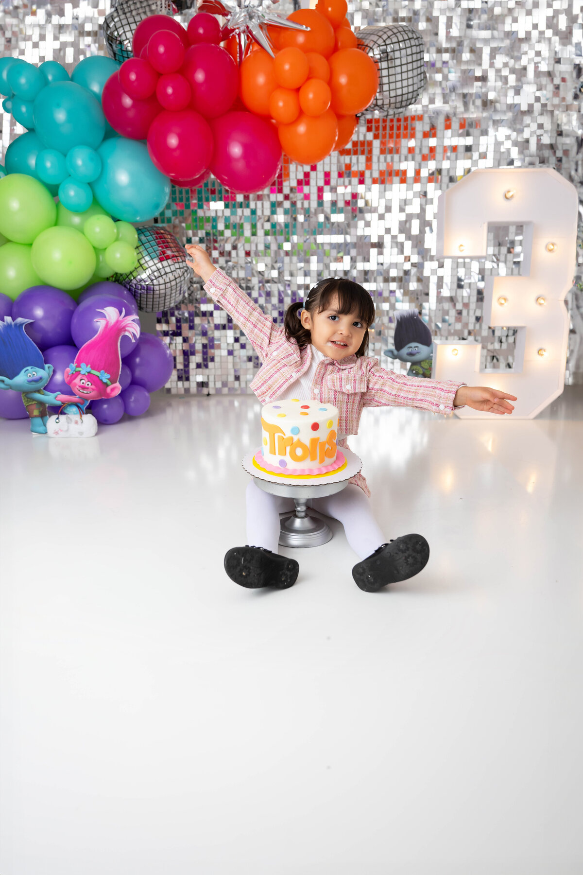 With hands stretched out, child sits with a cake between her legs wearing a plaid pink and white outfit. 
