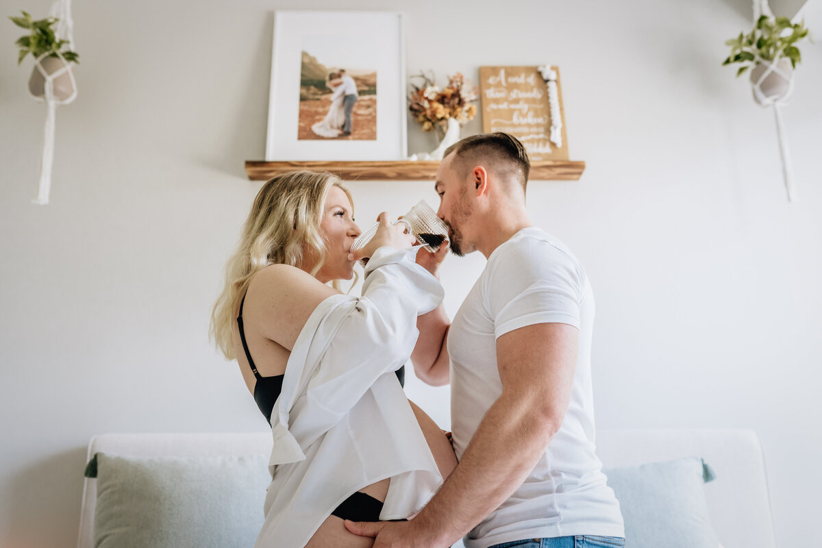 The couple stands facing each other in a bedroom, taking a sip from their mugs while holding each other, with a shelf displaying a wedding photo and a sign in the background.