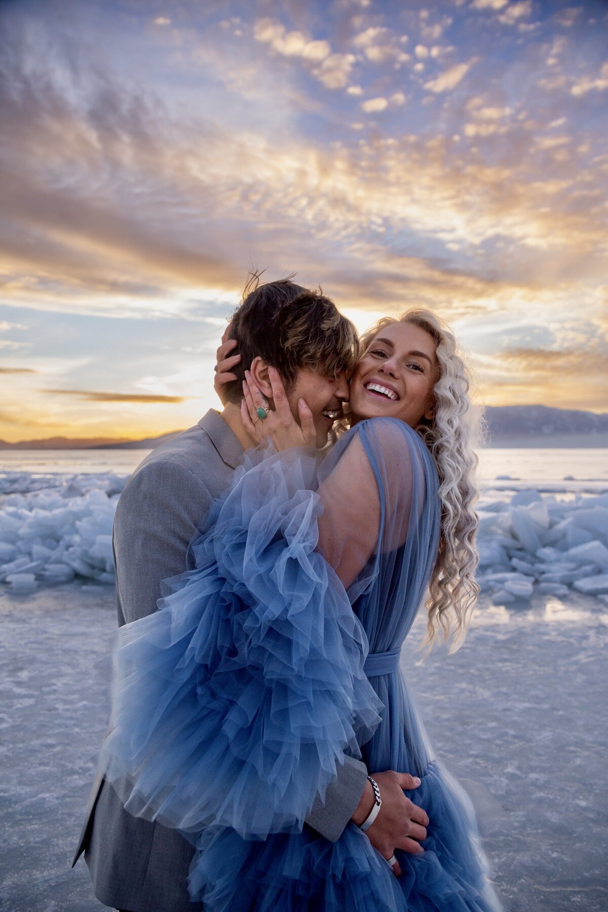 Groom embracing his bride and resting his face on the side of her face. Bride is looking at the camera with a big smile as she embraces her groom's face with both hands. They are standing in front of the ice stacks at sunset at Utah Lake.
