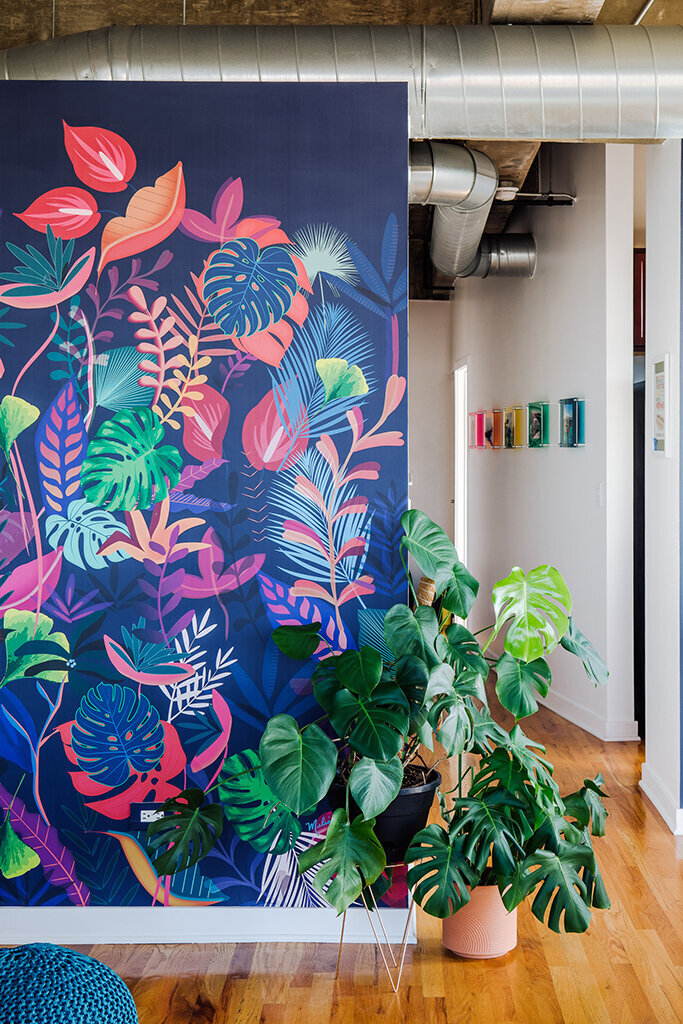 A vibrant and colorful tropical wall mural dominates the left side of the frame, featuring large stylized leaves in shades of blue, pink, green, and orange against a dark blue background. Two large potted monstera plants stand in front of the mural on a wooden floor. Exposed silver ductwork runs along the ceiling. A glimpse into another room shows a white wall with colorful framed artwork.
