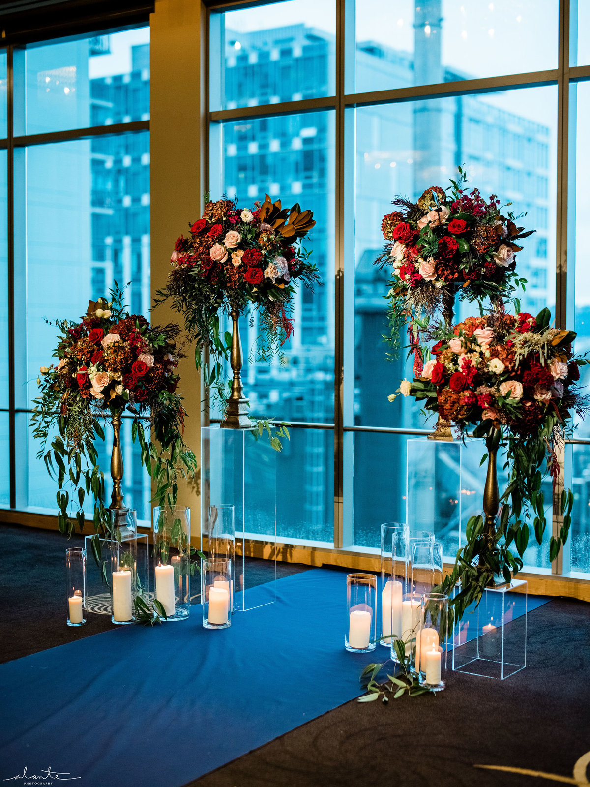 Wedding ceremony flowers in red and greens, with navy blue aisle runner, and hurricane candles