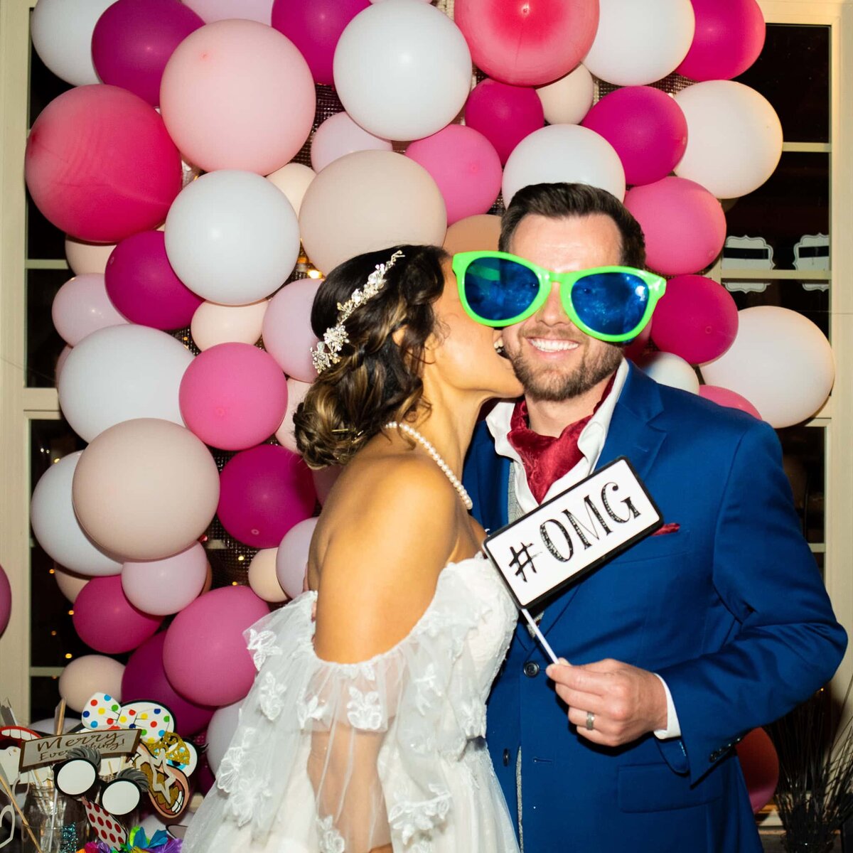 Groom having fun at a photo booth with oversized sunglasses and a sign that says OMG while his bride bites his cheek in front of many pink balloons.