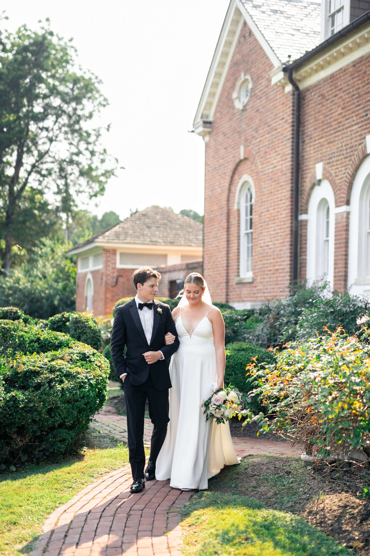 A bride and groom walk arm in arm on a brick path beside a red brick building. The bride wears a white dress and holds a bouquet, while the groom wears a black suit. The scene is bright and surrounded by greenery.