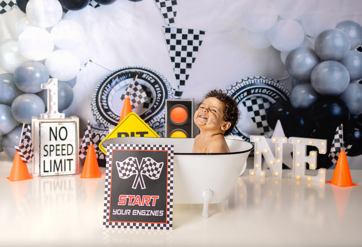 Smiling baby boy in race car themed cake smash bath setup with start your engines sign during first birthday session in Waldwick NJ