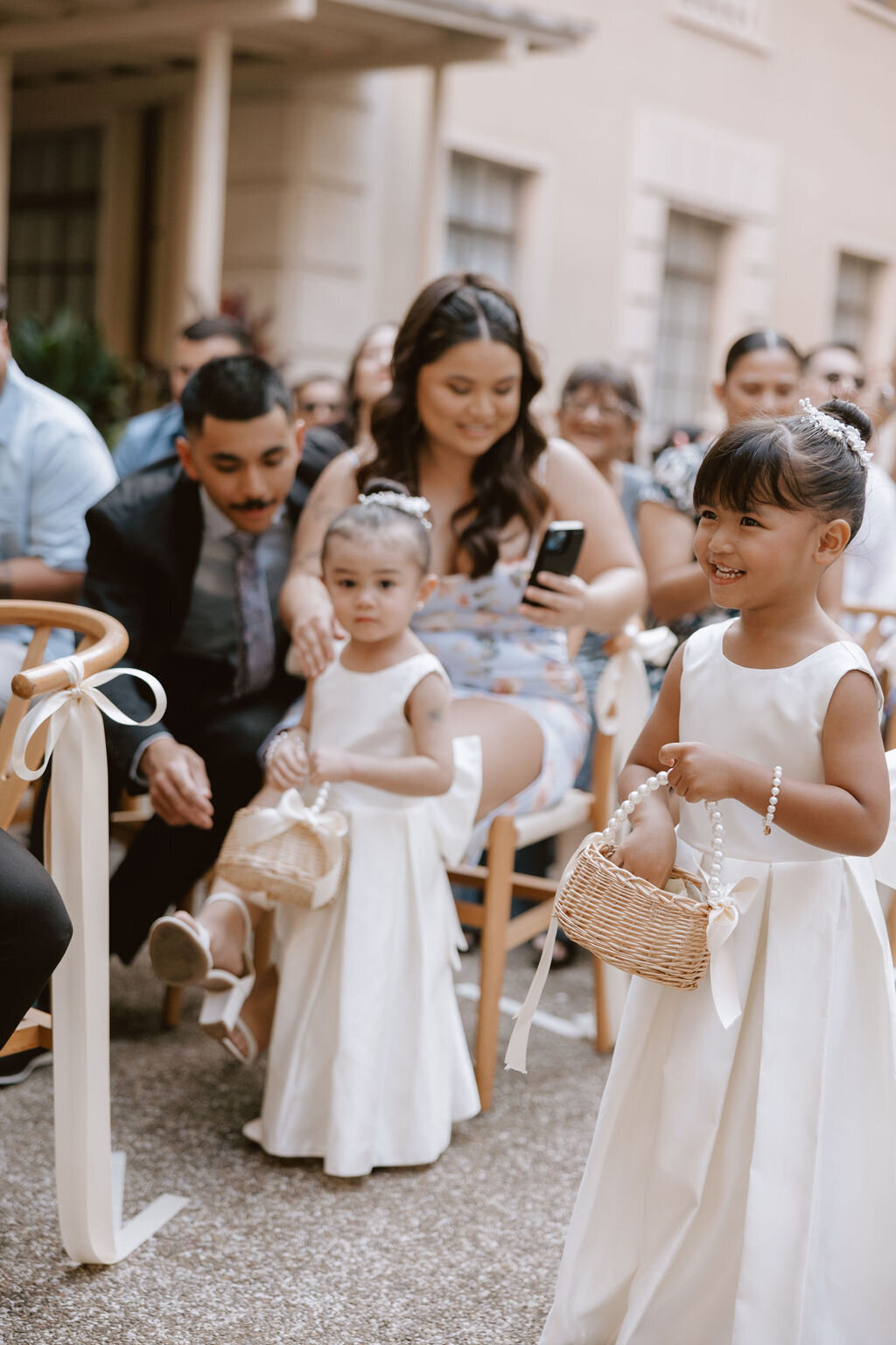 Flower girls walking down the aisle in ivory dresses while mom takes phone photo in the background seated