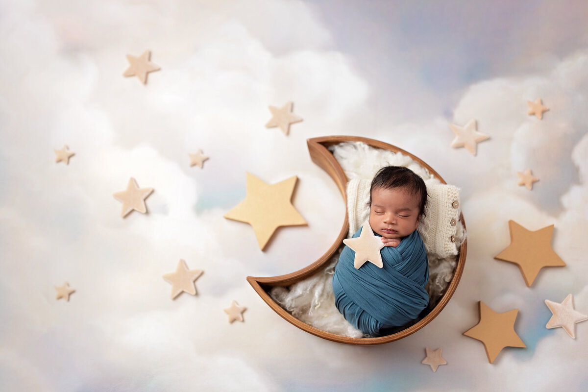 Newborn baby sleeping in a crescent bowl surrounded by clouds and gold stars.