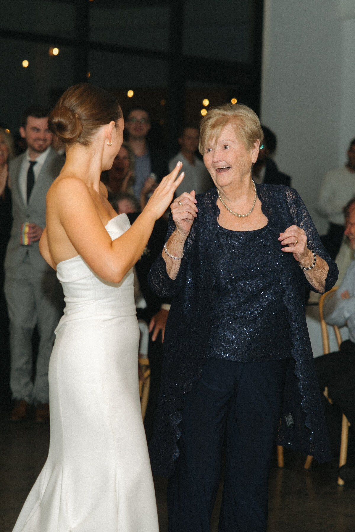 A bride in a formal, fitted, white, strapless wedding dress dances with her right arm bent upward. She faces her grandma with a navy blue pant with a blue shawl. Her grandma smiles and snaps her fingers dancing and looking at the bride. Wedding guests stand and sit behind them watching them dance. 
