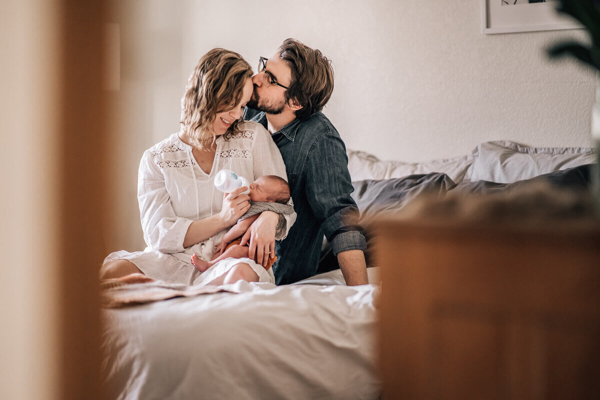 Minneapolis newborn photographer, Kate Simpson, takes a pull back shot through the doorway of a mom feeding her baby while Dad kisses Mom's forehead.