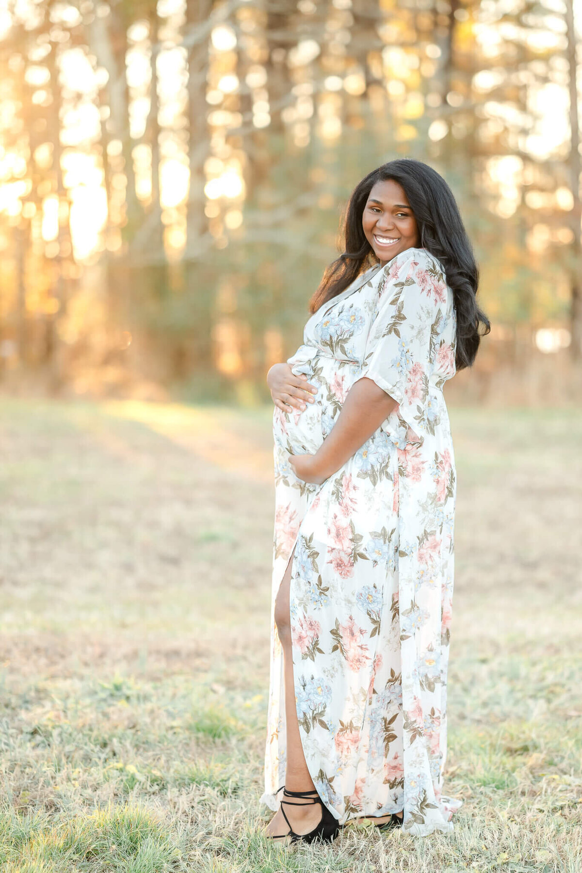 A pregnant woman, in a white floral dress, smiles during her Chesapeake Maternity session. She is holding her belly and the sun is glowing behind her.