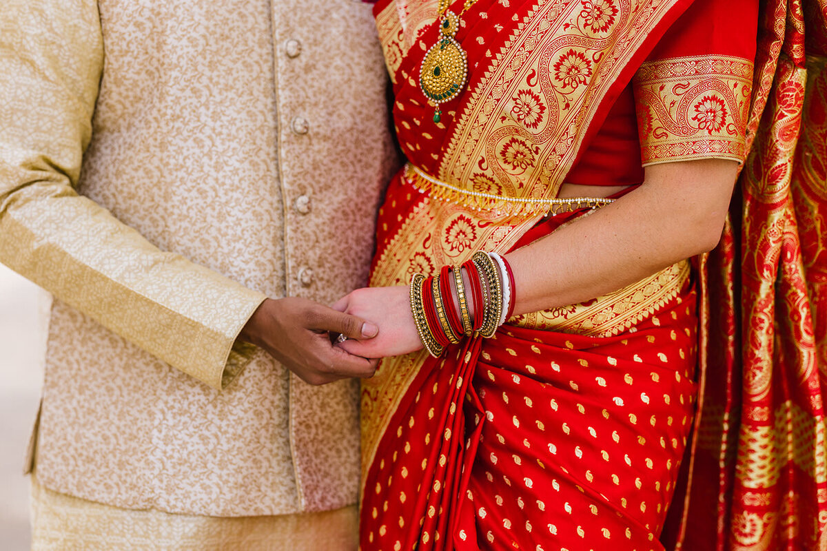 A detail shot of a bride and groom's hands as they hold each other, showcasing the bride's red saree, bangles, and henna.