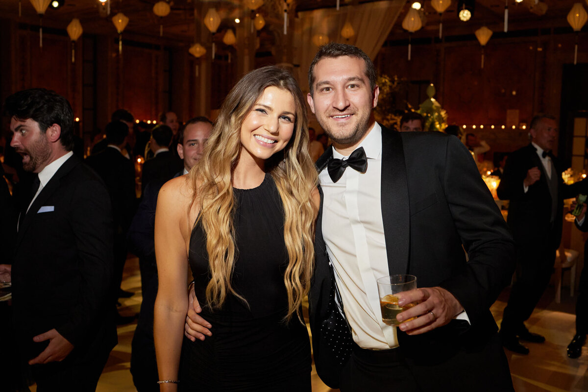 The woman and man, dressed formally, are seen at an indoor event at The New York Public Library in New York City. The woman, in a black sleeveless dress, is captured smiling widely while the man, sporting a black tuxedo and bow tie, holds a drink in his hand. The background features other attendees and decorative elements, providing a vibrant setting for the occasion. The photo was taken by photographer Shawn Connell, adding a professional touch to the elegant scene. Additionally, Aaron Navak Films provided videography services, capturing the event in stunning detail.