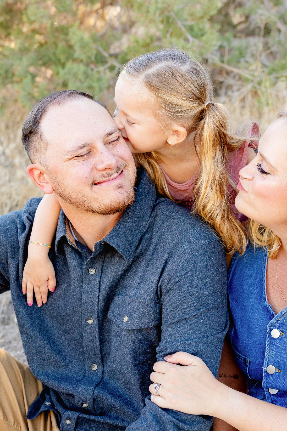 A father smiles as his daughter kisses his cheek while the mother looks on playfully. They are embracing closely, sharing a warm and loving moment with a natural outdoor background.