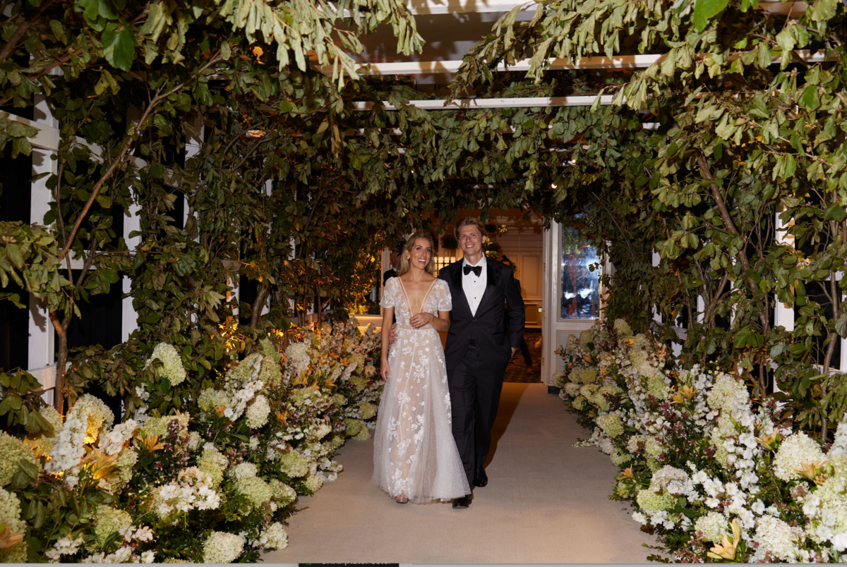 This is where the photo was taken at Washington D.C., The Willard Intercontinental, top wedding venues and hotels in DC by photographer Shawn Connell. In the scene, a couple walks arm in arm down an aisle adorned with lush greenery and white flowers. The woman is wearing a white floral-patterned gown, and the man is dressed in a black tuxedo. They are smiling and appear to be at a formal event or wedding.