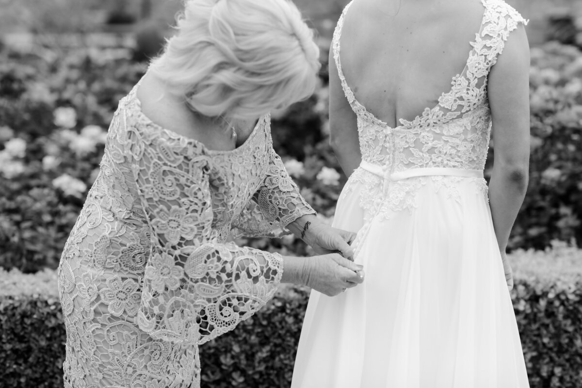 brides mother buttons up her daughters wedding dress while they stand together in a rose garden for little rock wedding