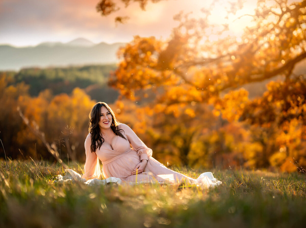 A mama to be laughs and cradles her belly while sitting on a blanket in the grass