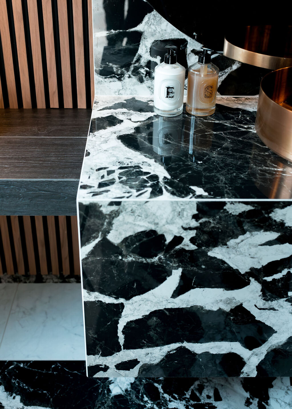 A close-up view of the marble countertop in a dark, modern bathroom. The side of a copper sink bowl is visible. To the left, there is a wooden slatted wall.